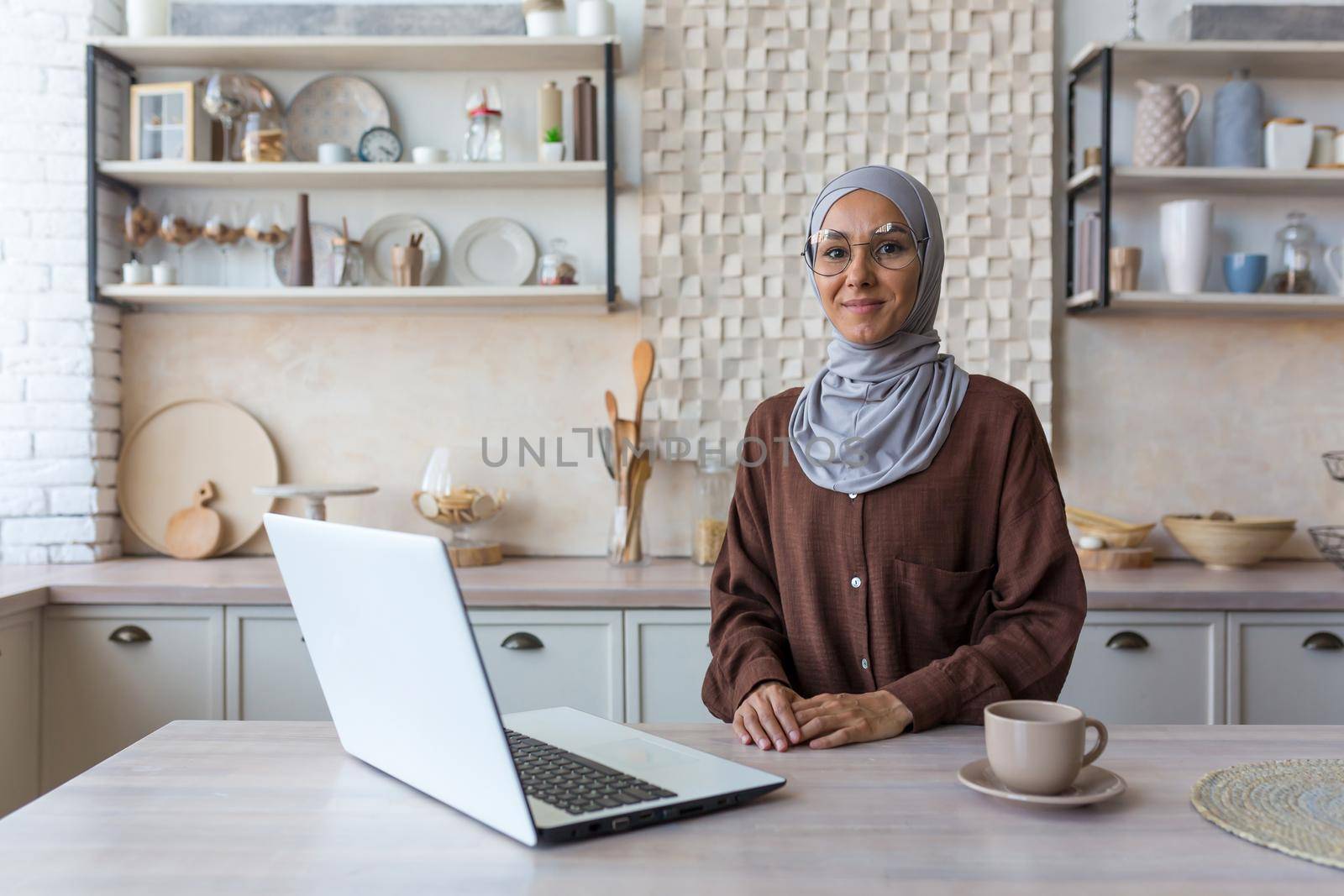 Portrait of young Muslim woman in hijab, woman in glasses looking at camera studying at home sitting in kitchen, female student using laptop for remote online learning.