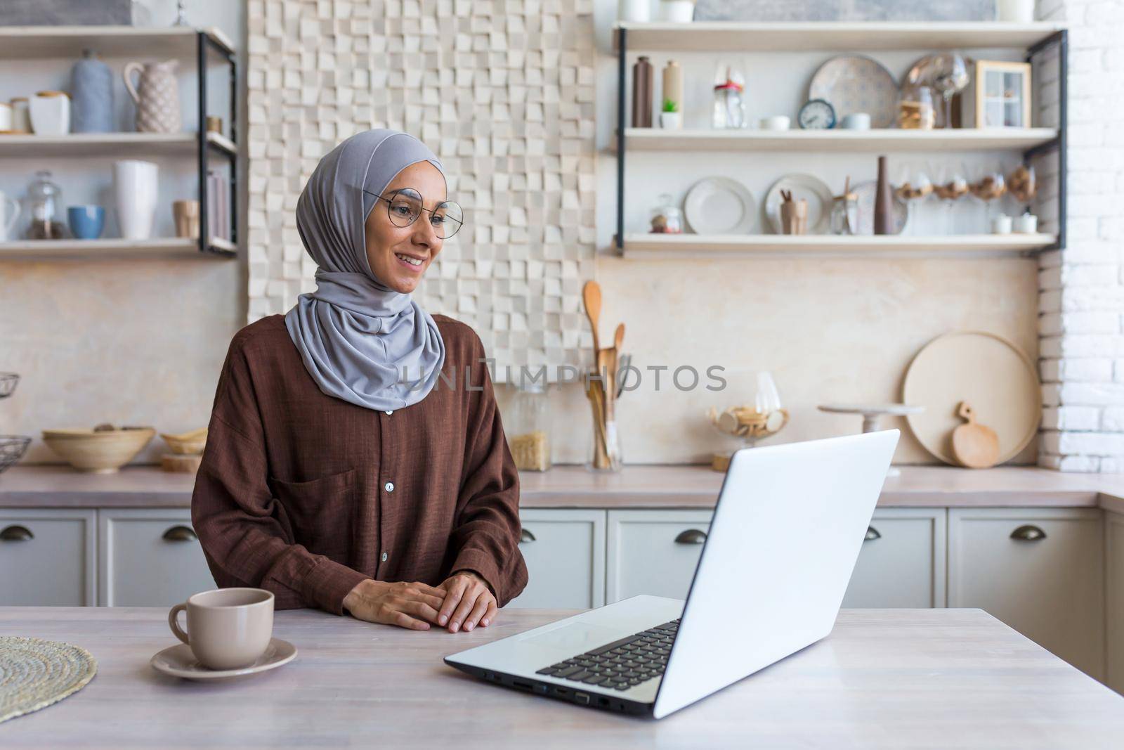 Young beautiful muslim woman in hijab and glasses studying remotely using laptop student woman in kitchen smiling and happy.