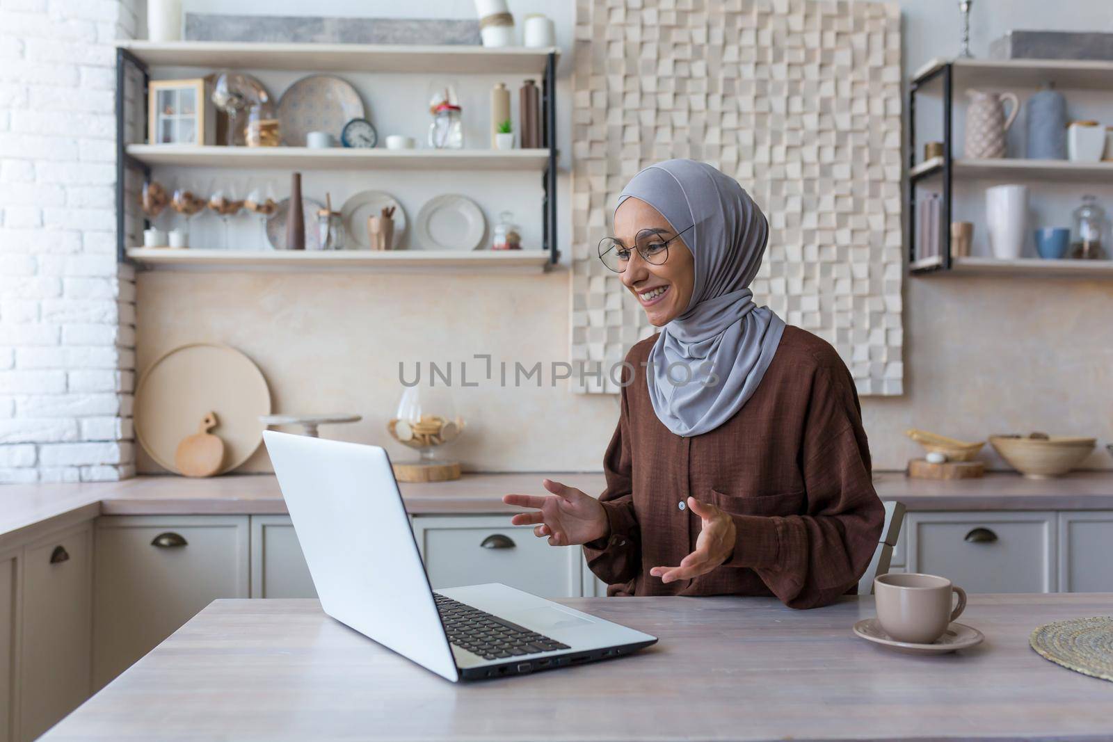 Young beautiful Muslim woman in hijab using laptop for video call, woman at home sitting in kitchen talking with friends remotely smiling at computer web camera
