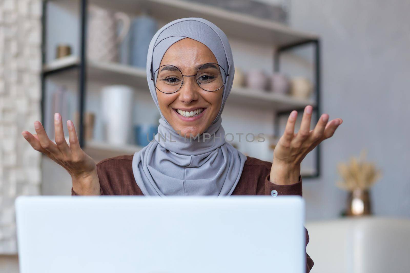 Video call online meeting with friends, young beautiful muslim woman in hijab looking at camera and smiling talking with friends remotely, woman at home in kitchen using laptop.
