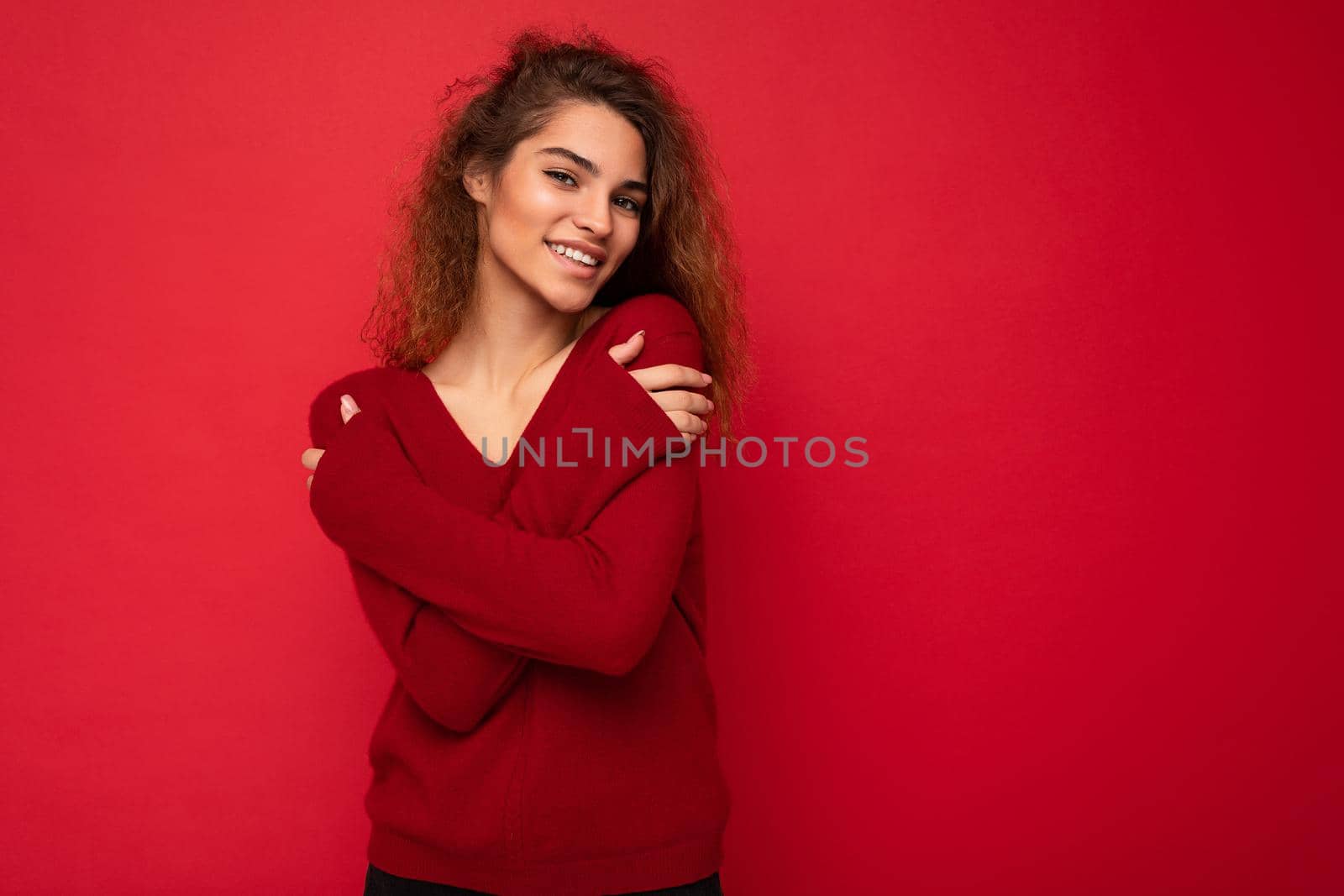 Photo of beautiful attractive charming positive happy smiling young brunette curly woman wearing stylish dark red sweater isolated over red background wall with free space for text.