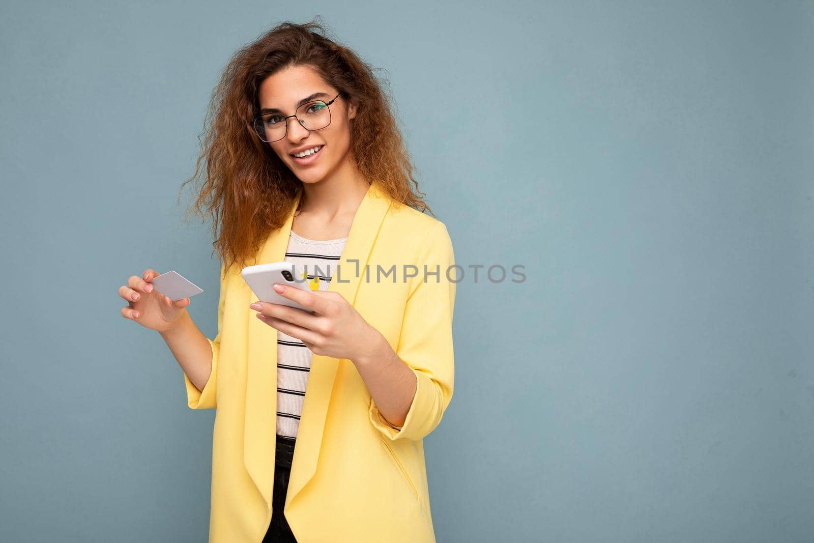 Portrait of attractive young woman with curly dark blond hair wearing yellow jacket and optical glasses isolated on background with free space holding and using phone looking at camera.
