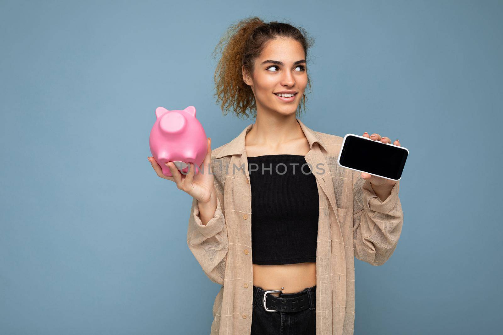 Portrait of happy positive smiling young beautiful attractive brunette curly woman with sincere emotions wearing stylish beige shirt isolated over blue background with copy space, holds pink pig money box and shows mobile phone with empty screen display. Payment concept.