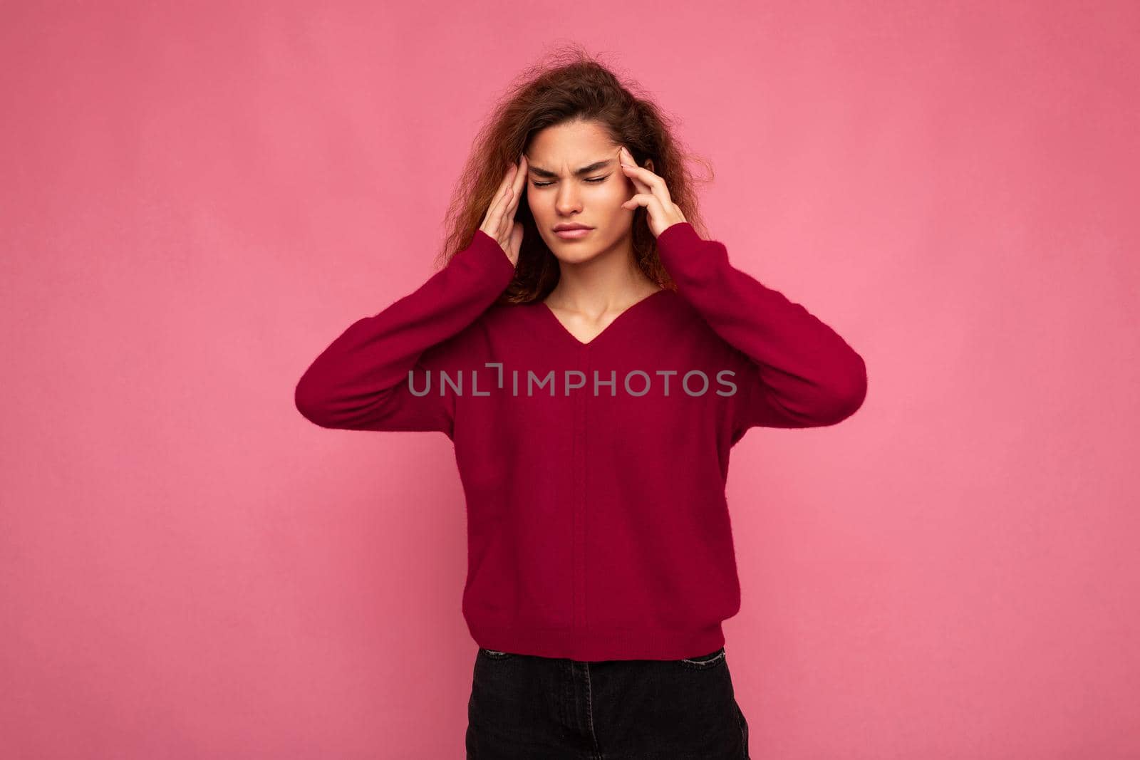 Portrait of young emotional beautiful brunette curly woman with sincere emotions wearing trendy pink pullover isolated on pink background with copy space and having headache.