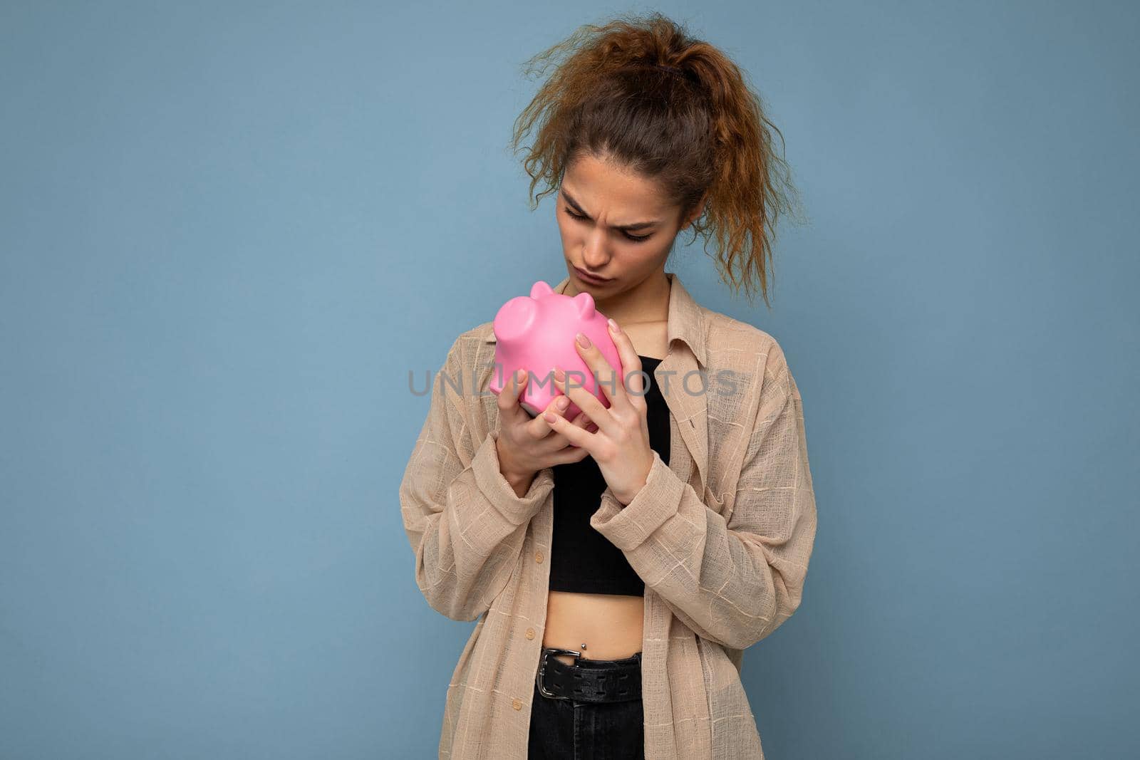 Portrait photo of sincere emotional cute young beautiful attractive woman with dark curly hair in a ponytail wearing casual beige shirt isolated over blue background with copy space and holds pink piggy box. Moneybox concept.