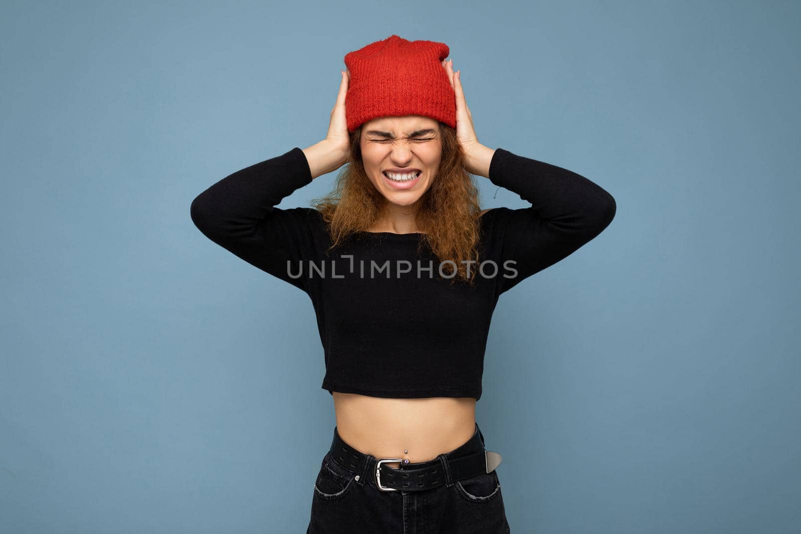 Shot of young emotional nice winsome brunet woman wavy-haired with sincere emotions wearing black crop top and red hat isolated on blue background with free space and covering ears.