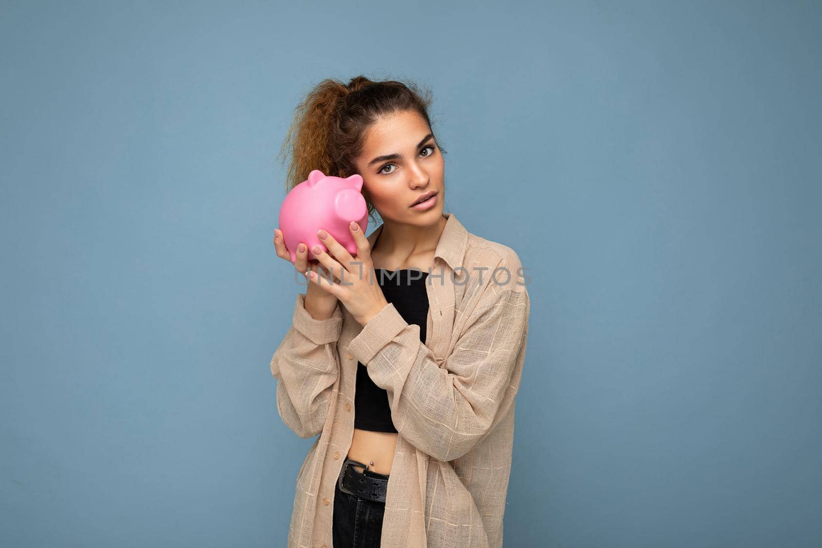 Portrait photo of sincere cute young beautiful attractive woman with dark curly hair in a ponytail wearing casual beige shirt isolated over blue background with copy space and holds pink piggy box. Moneybox concept.