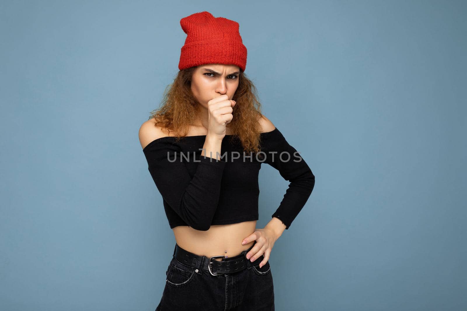 Shot of young emotional angry nice winsome brunet woman wavy-haired with sincere emotions wearing black crop top and red hat isolated on blue background with free space and holding hand near mouth.