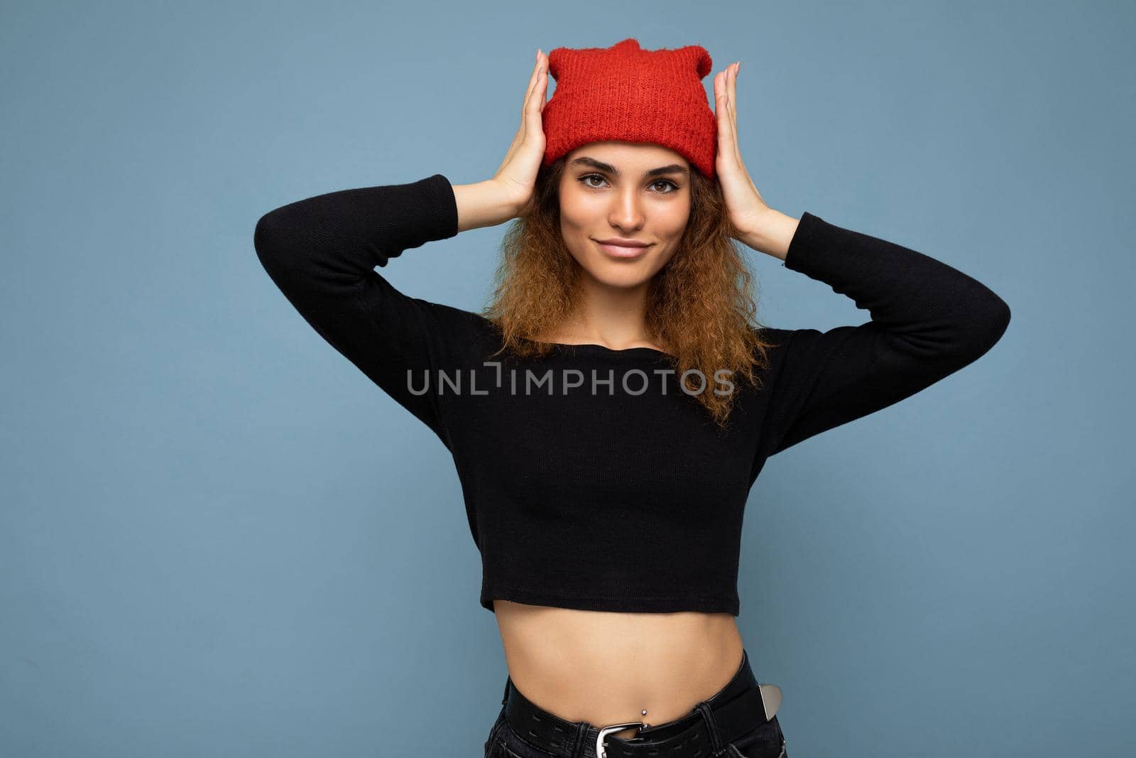 Photo of young pretty positive brunette woman curly with sincere emotions wearing stylish black crop top and red hat isolated on blue background with copy space.