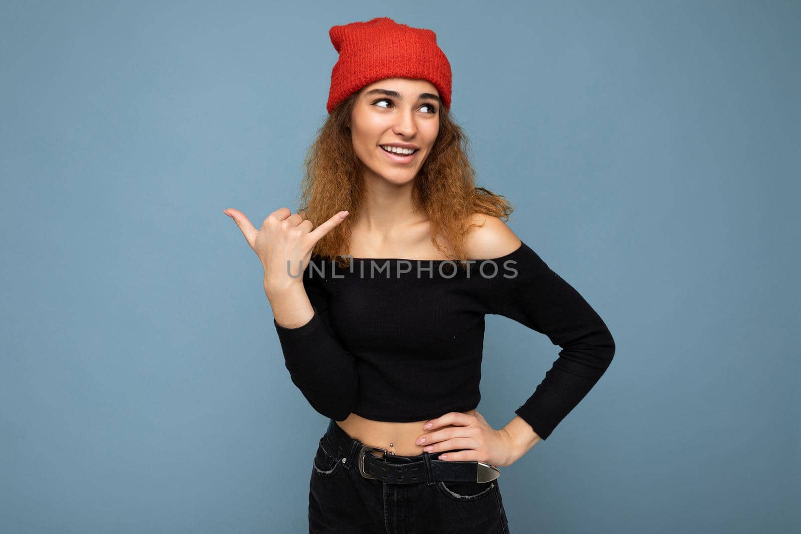 Photo of young positive happy beautiful brunette curly woman with sincere emotions wearing black crop top and red hat isolated on blue background with copy space and showing smartphone gesture with hand.