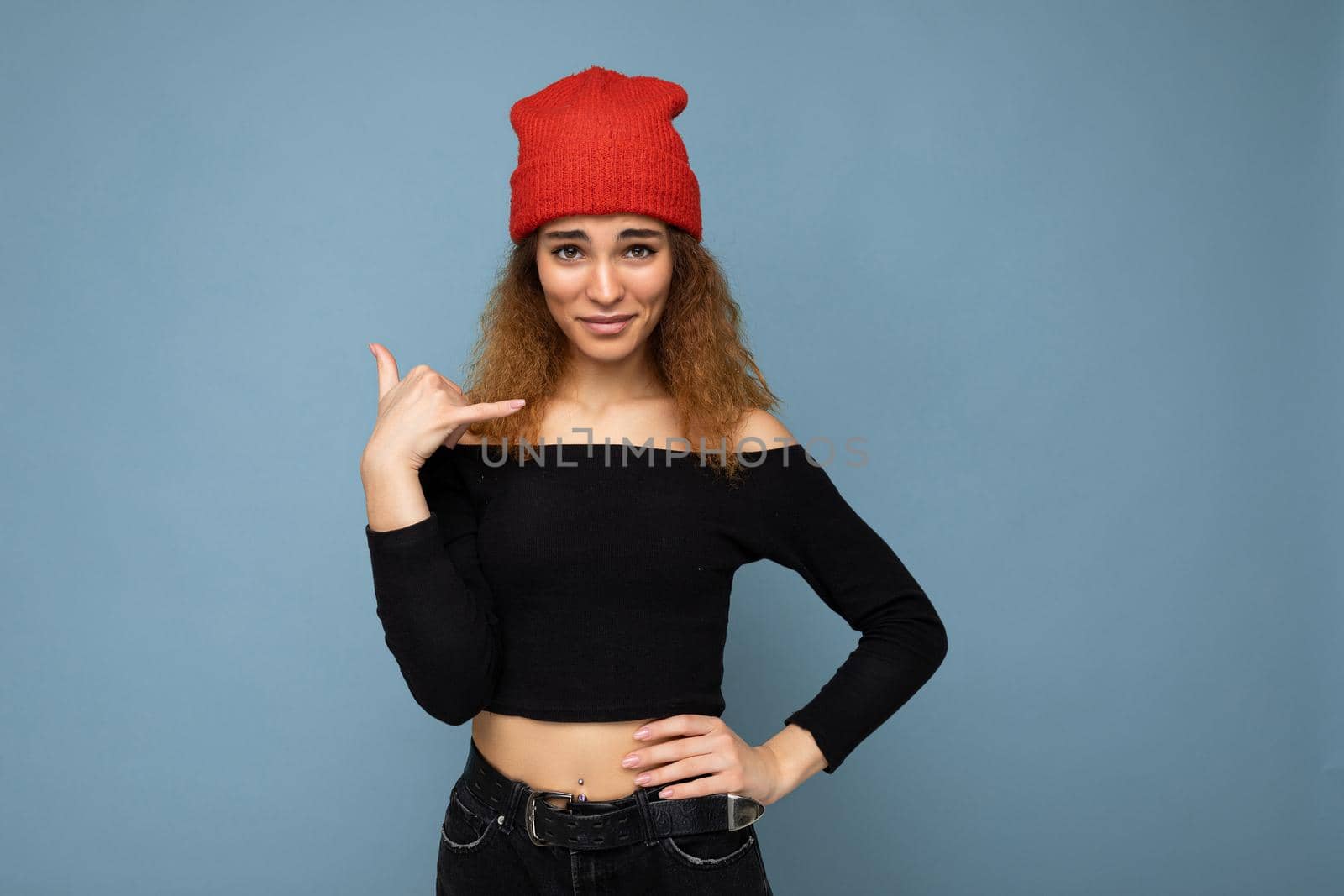 Photo of young positive beautiful brunette curly woman with sincere emotions wearing black crop top and red hat isolated on blue background with copy space and showing phone gesture.