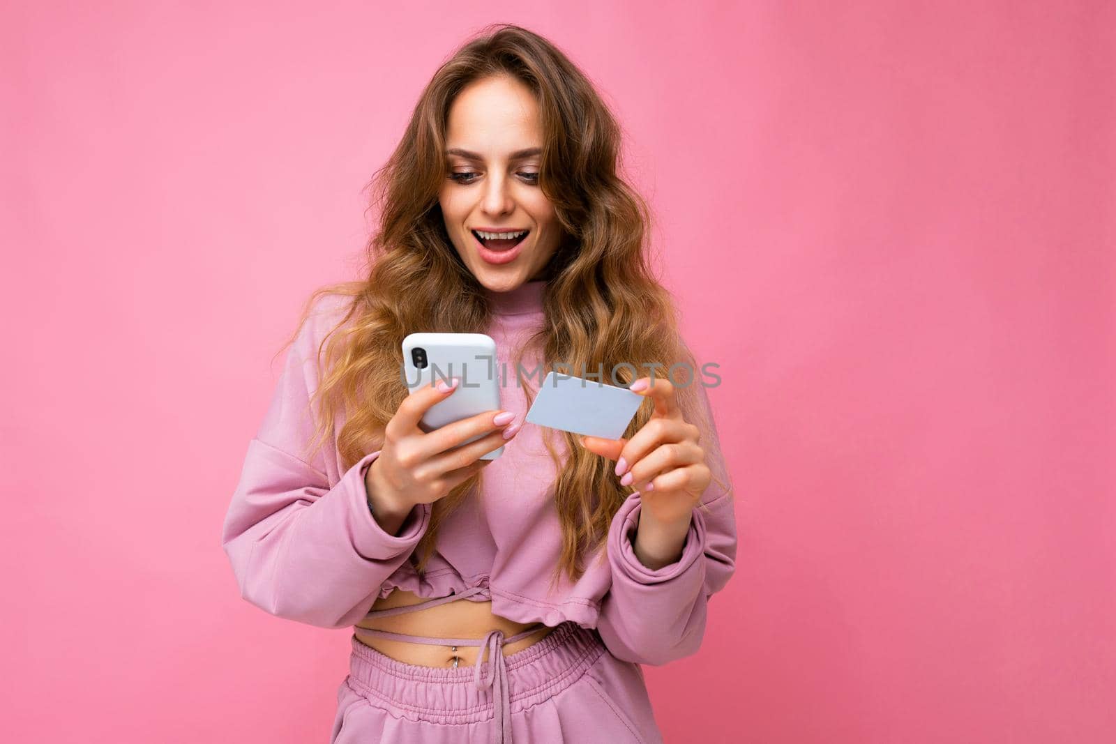 Photo shot of young beautiful attractive cute surprised happy curly blonde woman wearing trendy and stylish pink sport suit isolated over pink background with copy space for text and holding white plastic bank card and texting sms on mobile phone.