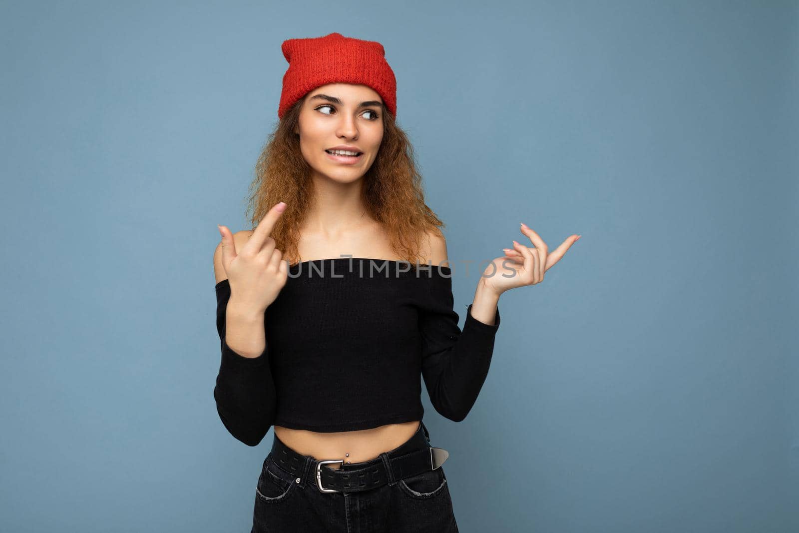 Photo of young positive attractive brunette curly woman with sincere emotions wearing black crop top and red hat isolated on blue background with empty space and pointig at free space.