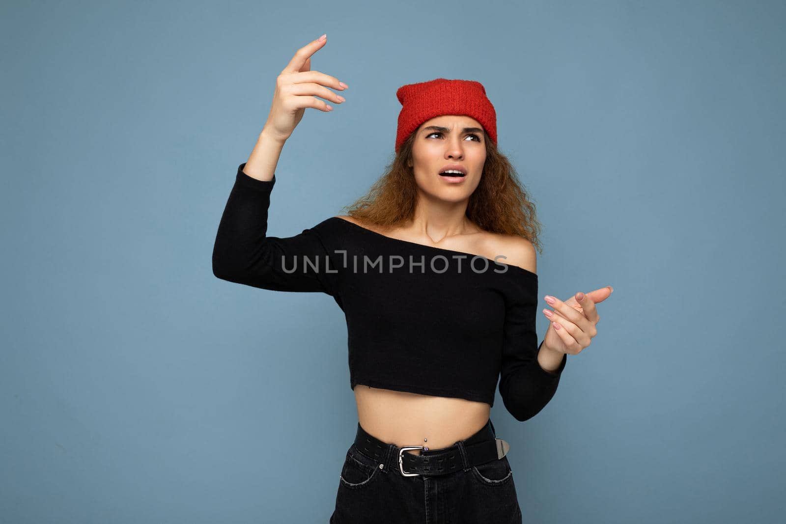 Young emotional dissatisfied pretty brunette woman wavy-haired with sincere emotions wearing black crop top and red hat isolated on blue background with free space.