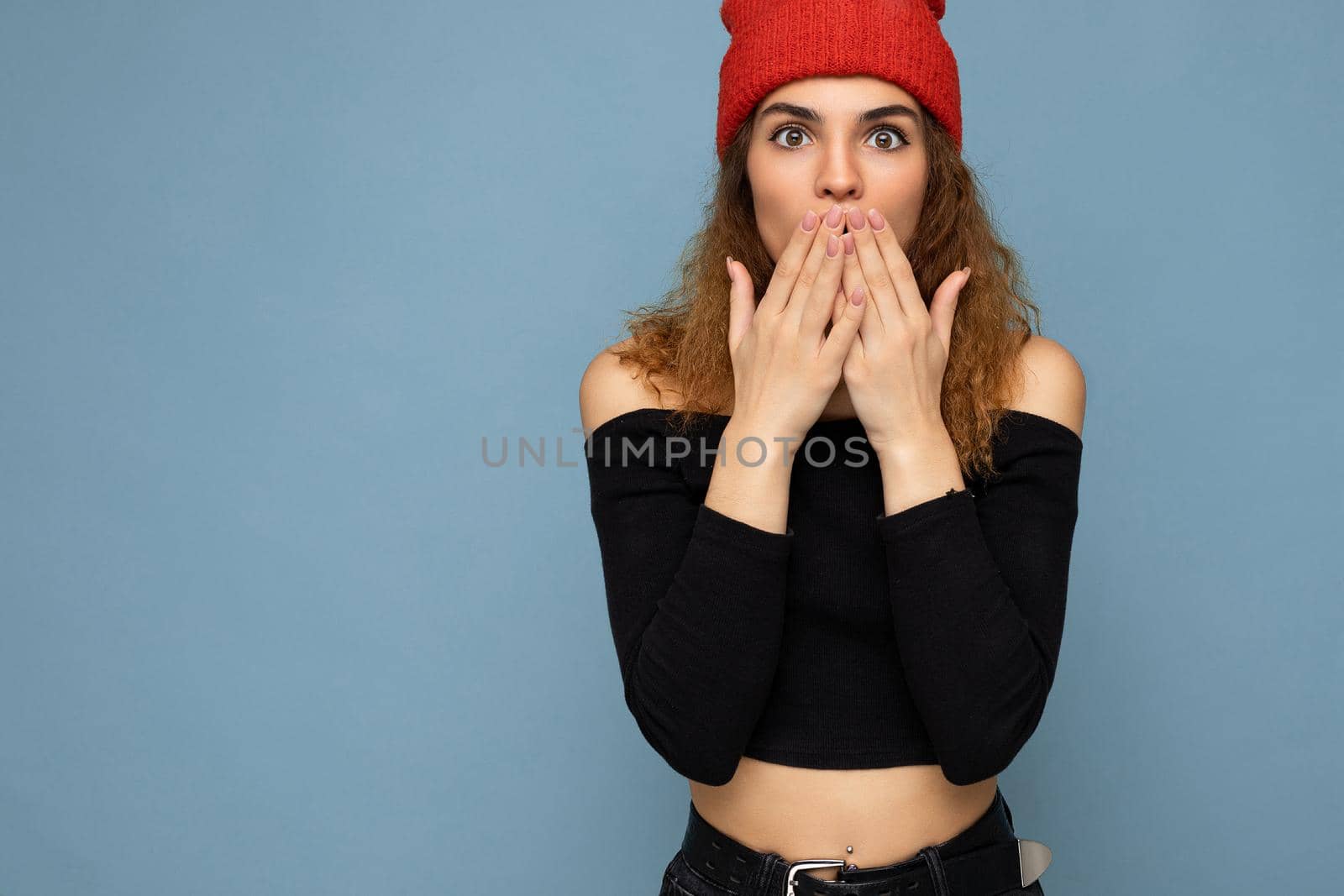 Shot of young emotional astonished nice winsome brunet woman wavy-haired with sincere emotions wearing black crop top and red hat isolated on blue background with free space and covering mouth.