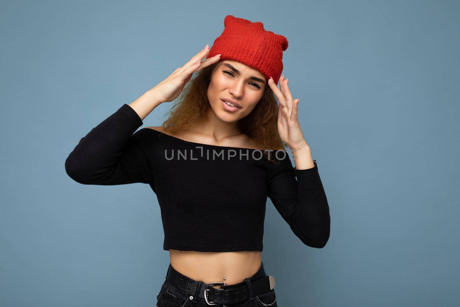 Young emotional dissatisfied pretty brunette woman wavy-haired with sincere emotions wearing black crop top and red hat isolated on blue background with free space and having headache.