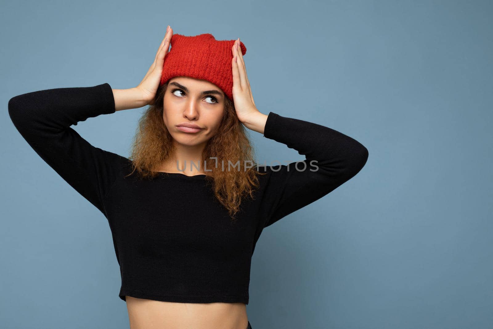 Photo of young pretty amusing positive brunette woman curly with sincere emotions wearing stylish black crop top and red hat isolated on blue background with copy space and grimacing.