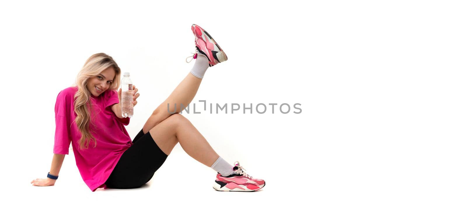 beautiful blonde sits against the background raising her legs in a tracksuit and shorts holding a bottle of water in her hands.