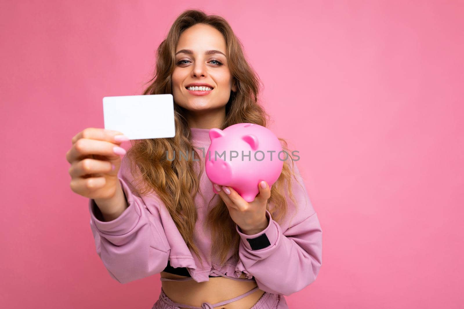 Photo shot of young beautiful attractive cute positive happy smiling curly blonde woman wearing trendy and stylish pink sport suit isolated over pink background with copy space for text and holding pink penny pig and white bank plastic card for money.