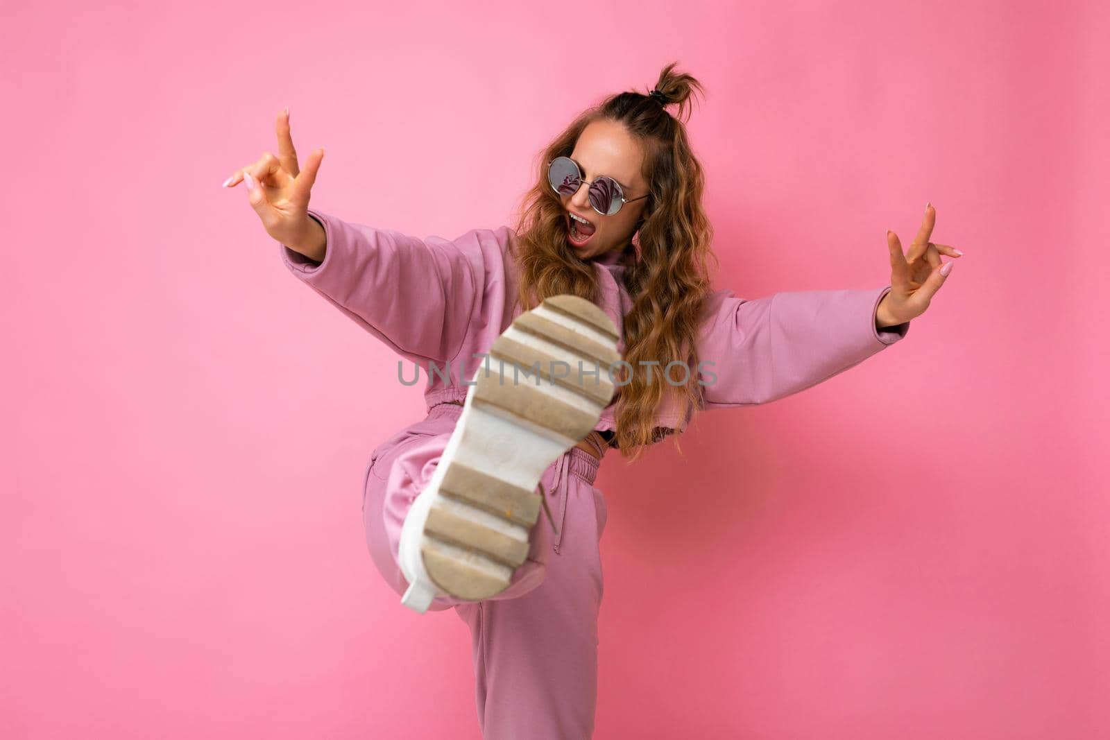 Photo shot of young beautiful attractive cute funny positive curly blonde woman wearing trendy and stylish pink sport suit and sunglasses isolated over pink background with copy space for text and having fun and joy.