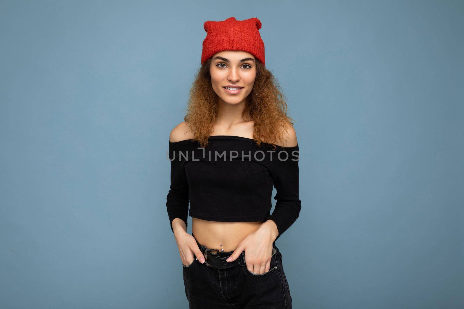 Photo of pretty positive smiling young brunet curly woman isolated over blue background wall wearing black crop top and stylish red hat looking at camera.