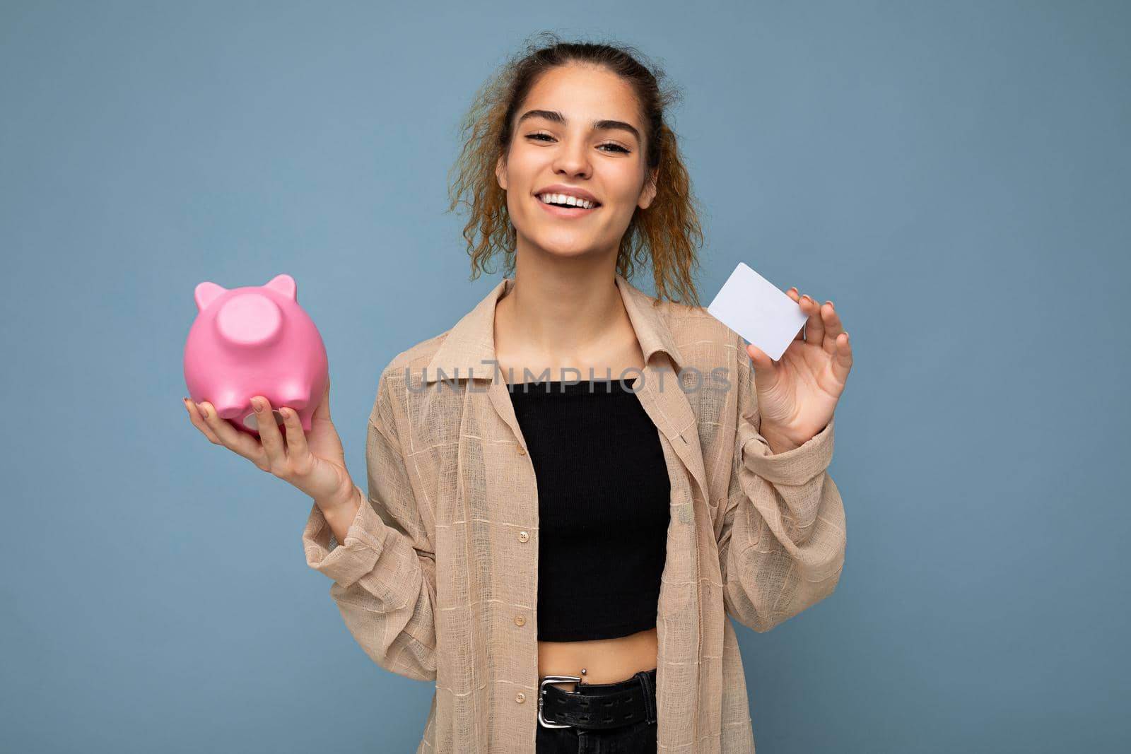 Portrait photo of happy positive smiling young beautiful attractive brunette curly woman with sincere emotions wearing stylish beige shirt isolated over blue background with copy space, holds pink piggy box and credit card. Shopping concept.