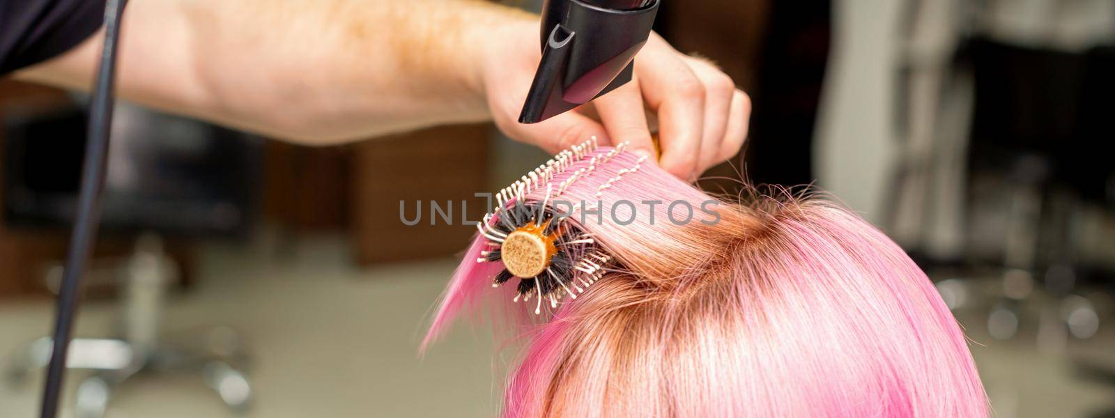 Drying short pink hair of young caucasian woman with a black hairdryer and black round brush by hands of a male hairdresser in a hair salon, close up