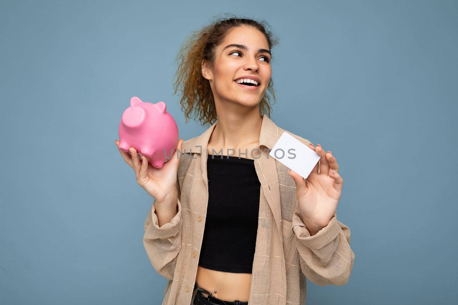 Portrait photo of happy positive smiling young beautiful attractive brunette curly woman with sincere emotions wearing stylish beige shirt isolated over blue background with copy space, holds pink pig penny bank and credit card. Shopping concept.