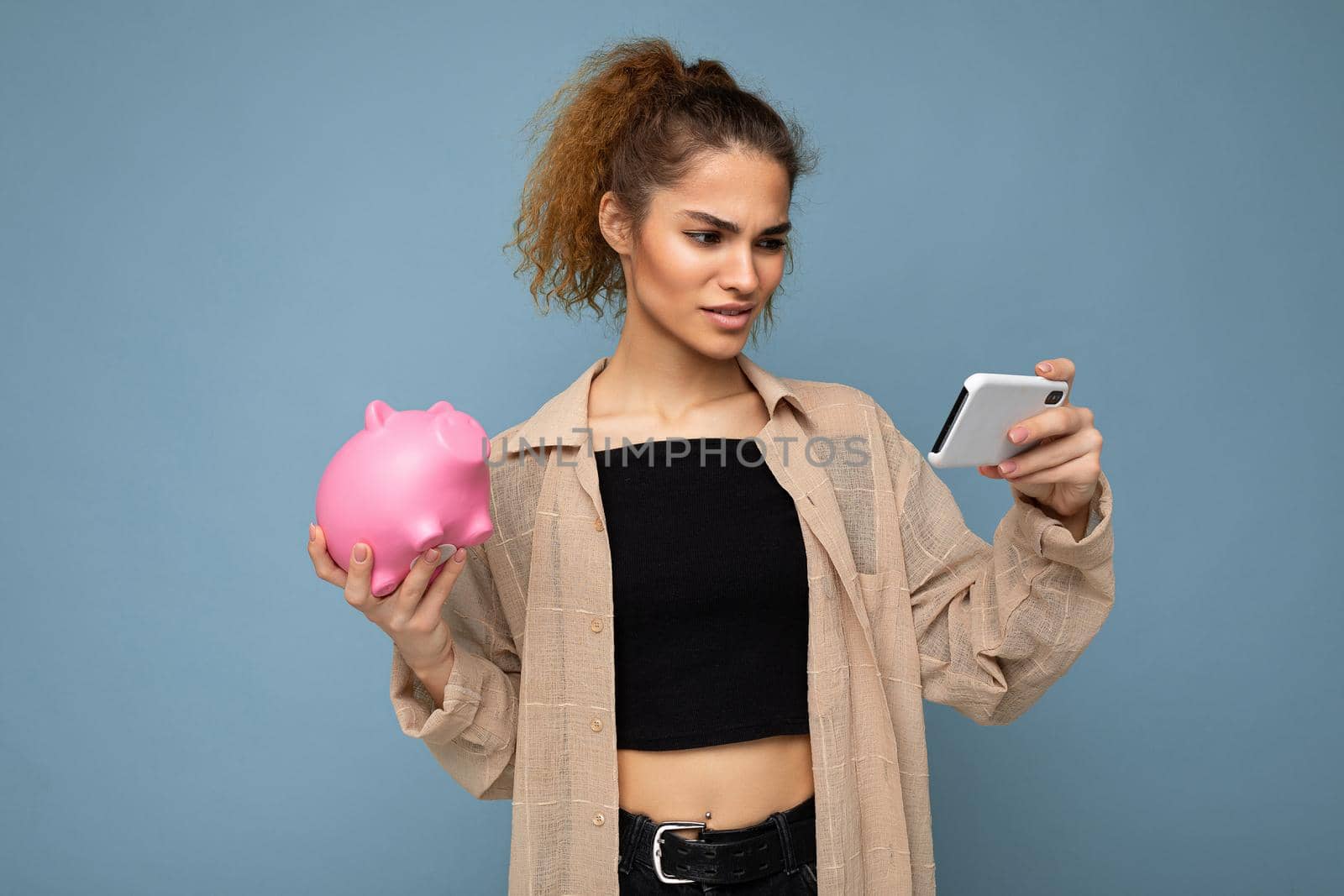 Portrait photo of dissatisfied young beautiful attractive brunette curly woman with sincere emotions wearing stylish beige shirt isolated over blue background with copy space, holds pink piggy box and uses mobile phone. I don't understand.
