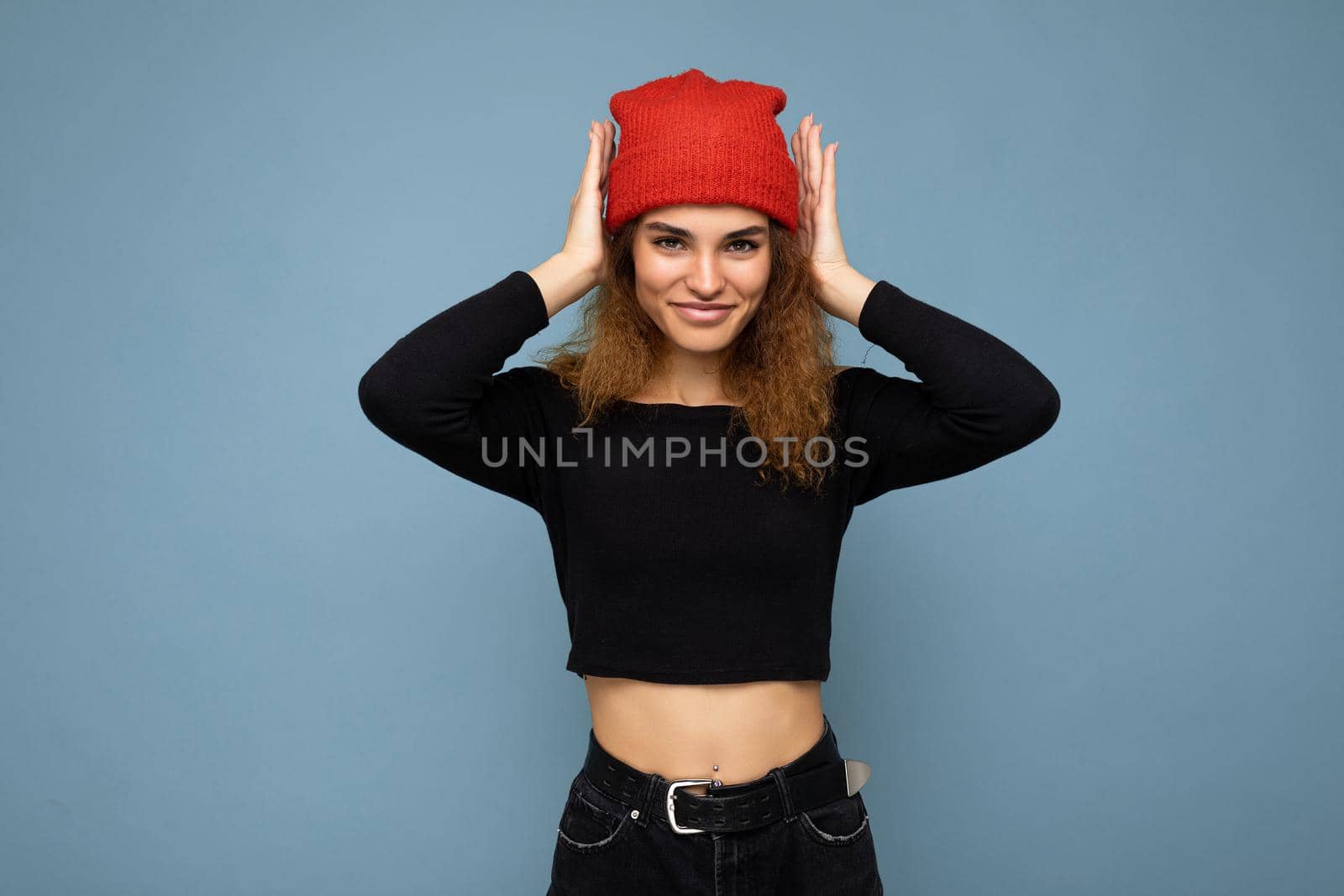 Shot of young nice winsome brunet woman wavy-haired with sincere emotions wearing black crop top and red hat isolated on blue background with free space and covering ears with hands.