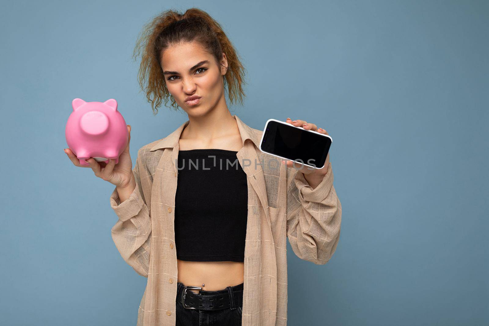 Photo of dissatisfied doubtful young beautiful attractive brunette curly woman with sincere emotions wearing stylish beige shirt isolated over blue background with copy space, holds pink pig penny bank for coins and shows smartphone with empty screen display. Payment concept.