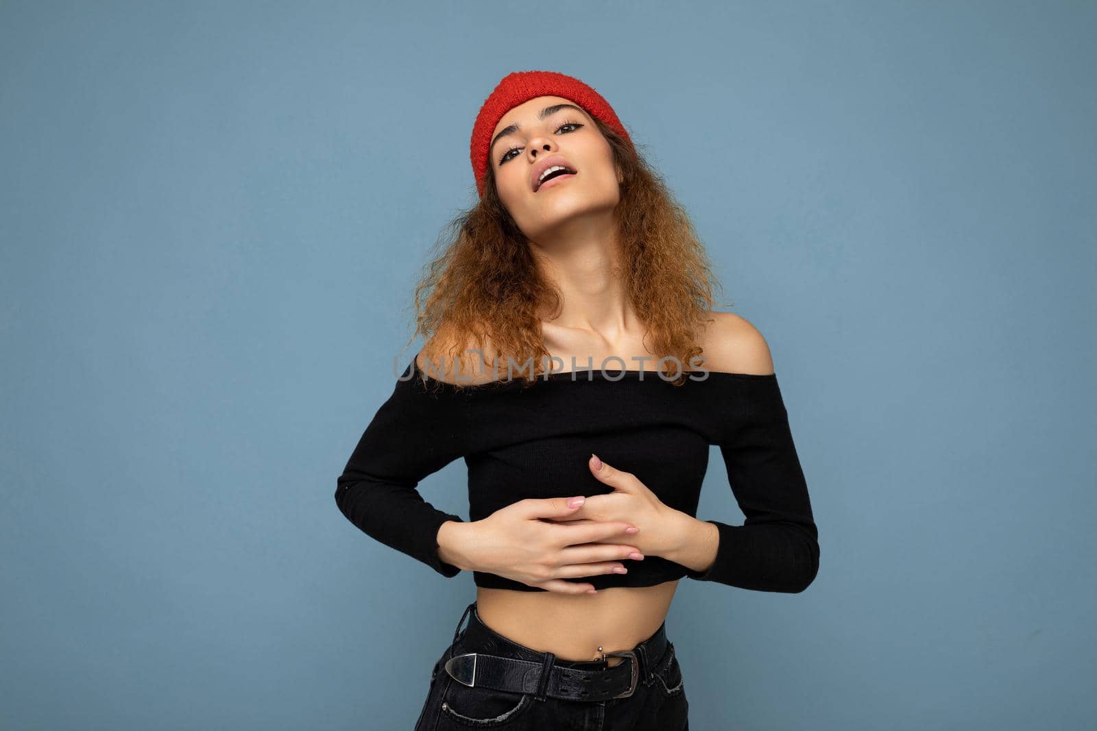 Photo of pretty beautiful attractive positive sexy cool young curly dark blonde woman standing isolated over blue background wall with copy space for text wearing everyday trendy and stylish black top and red hat and touching herself with hands.