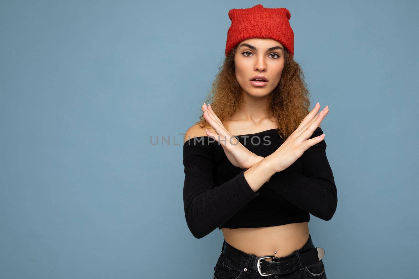 Shot of young amazed nice winsome brunet woman wavy-haired with sincere emotions wearing black crop top and red hat isolated on blue background with free space and showing stop gesture. Negative concept.
