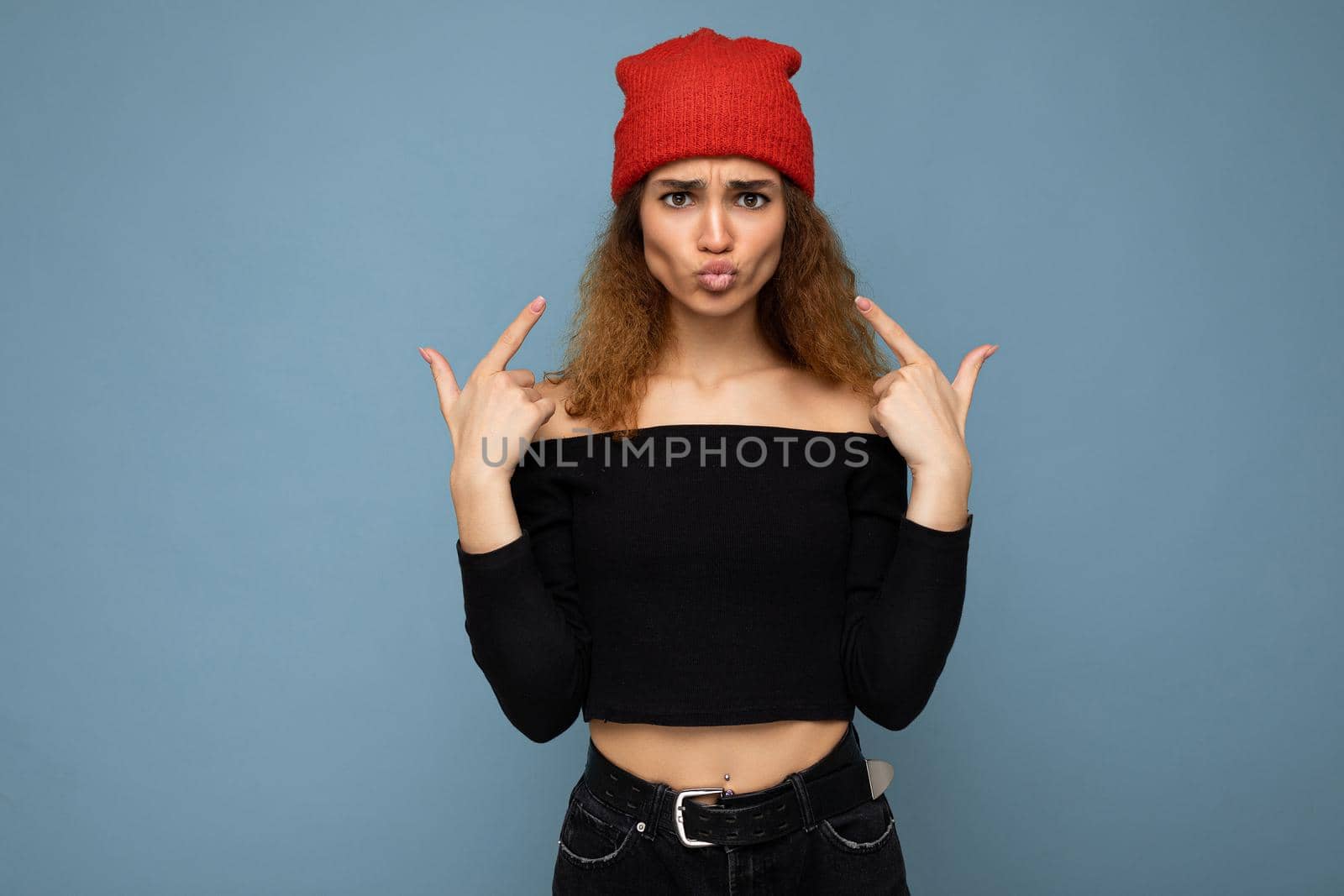 Photo of young positive beautiful brunette curly woman with sincere emotions wearing black crop top and red hat isolated on blue background with copy space and having fun.