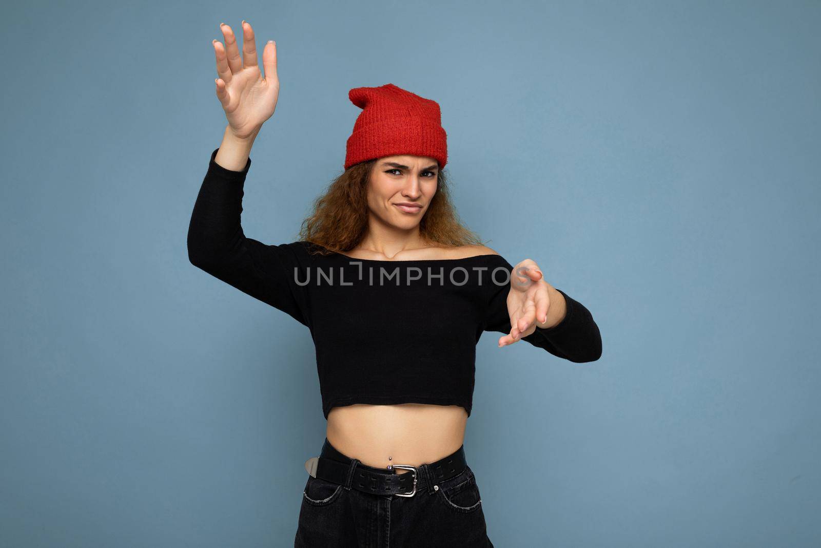 Photo of pretty beautiful attractive cool angry asking dissatisfied young curly dark blonde woman standing isolated over blue background wall with copy space for text wearing everyday trendy and stylish black top and red hat.