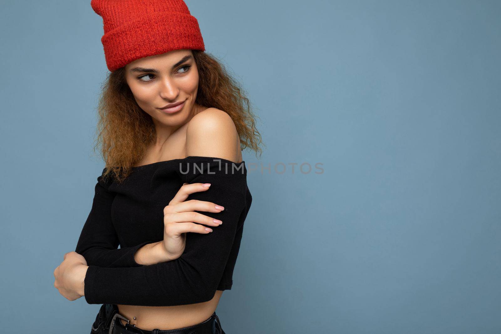 Photo of young positive sexy charming beautiful brunette woman curly with sincere emotions wearing stylish black crop top and red hat isolated on blue background with copy space and looking to the side.