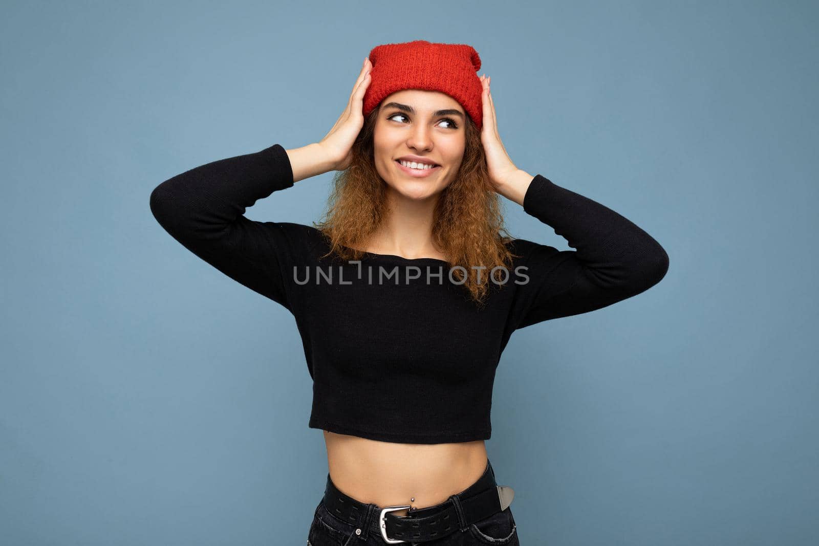 Photo of young positive smiling cute nice brunette woman curly with sincere emotions wearing stylish black crop top and red hat isolated on blue background with copy space by TRMK