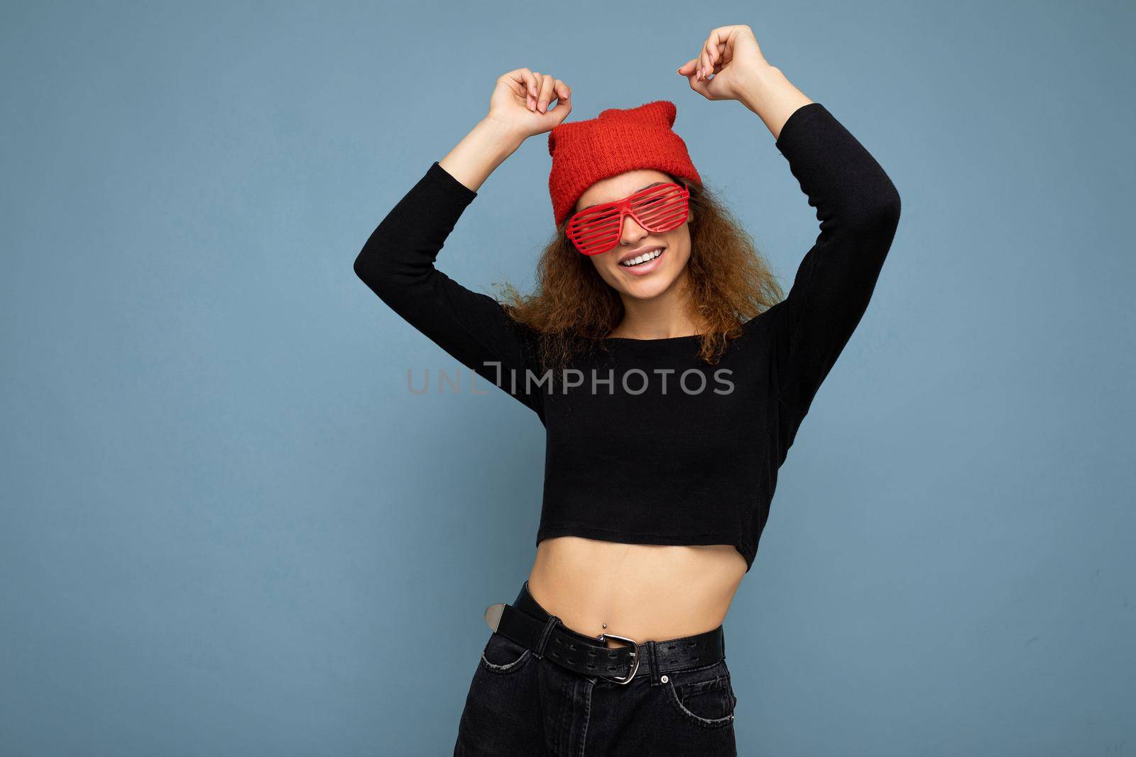 Photo of pretty beautiful attractive cool young curly dark blonde woman standing isolated over blue background wall with copy space for text wearing everyday trendy and stylish black top and red hat and glasses. Dancing and having fun.