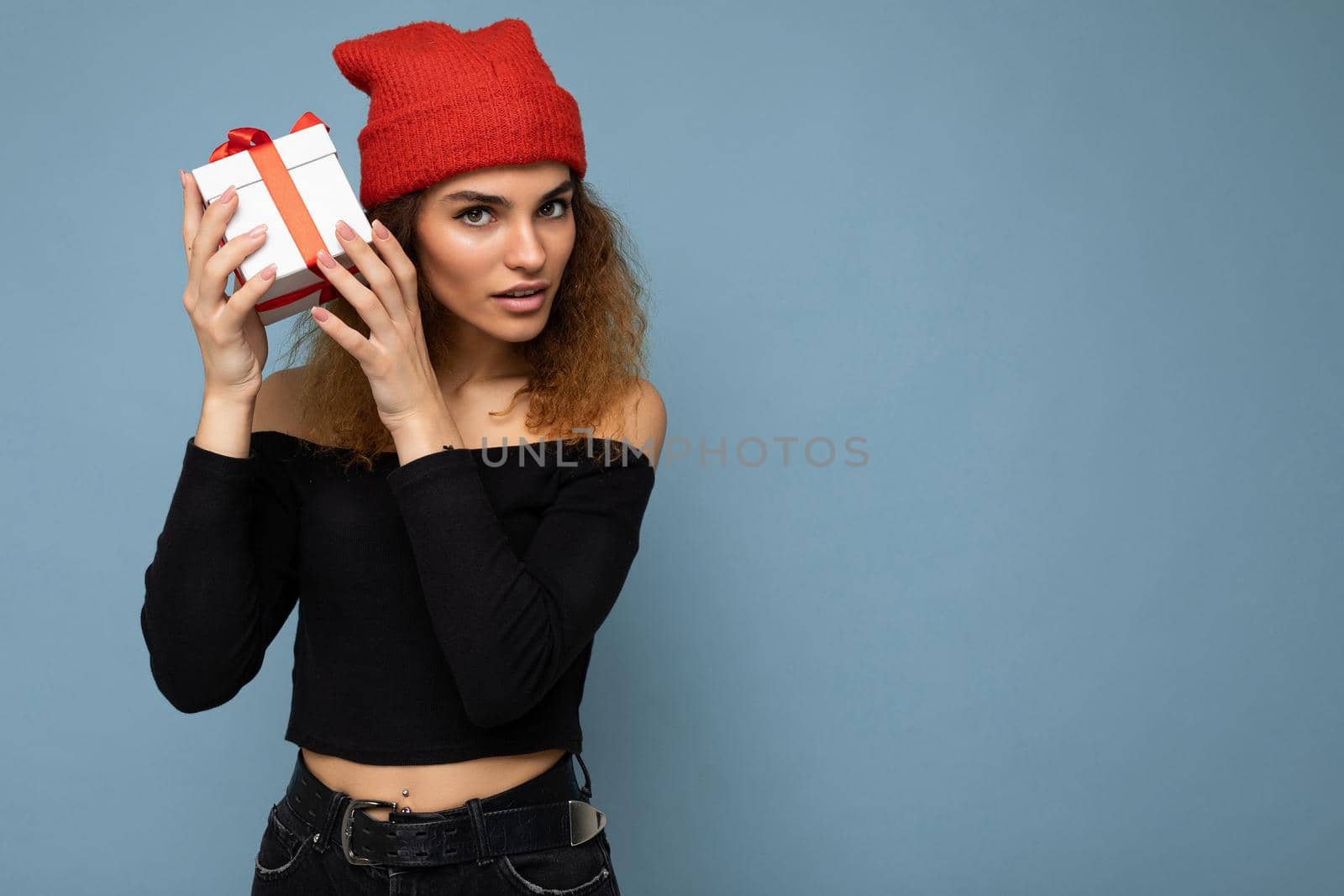 Photo shot of beautiful concentrated thinking adult dark blonde curly woman isolated over blue background wall wearing black crop top and red hat holding gift box looking at camera. Copy space, mockup