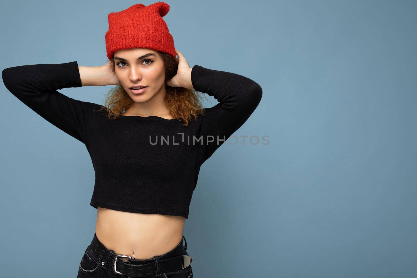 Photo of young positive cute nice brunette woman curly with sincere emotions wearing stylish black crop top and red hat isolated on blue background with copy space.