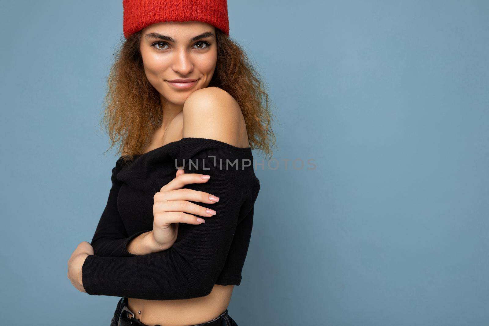 Photo of beautiful self-confident sexy young brunette curly woman isolated over blue background wall wearing black crop top and red hat looking at camera.