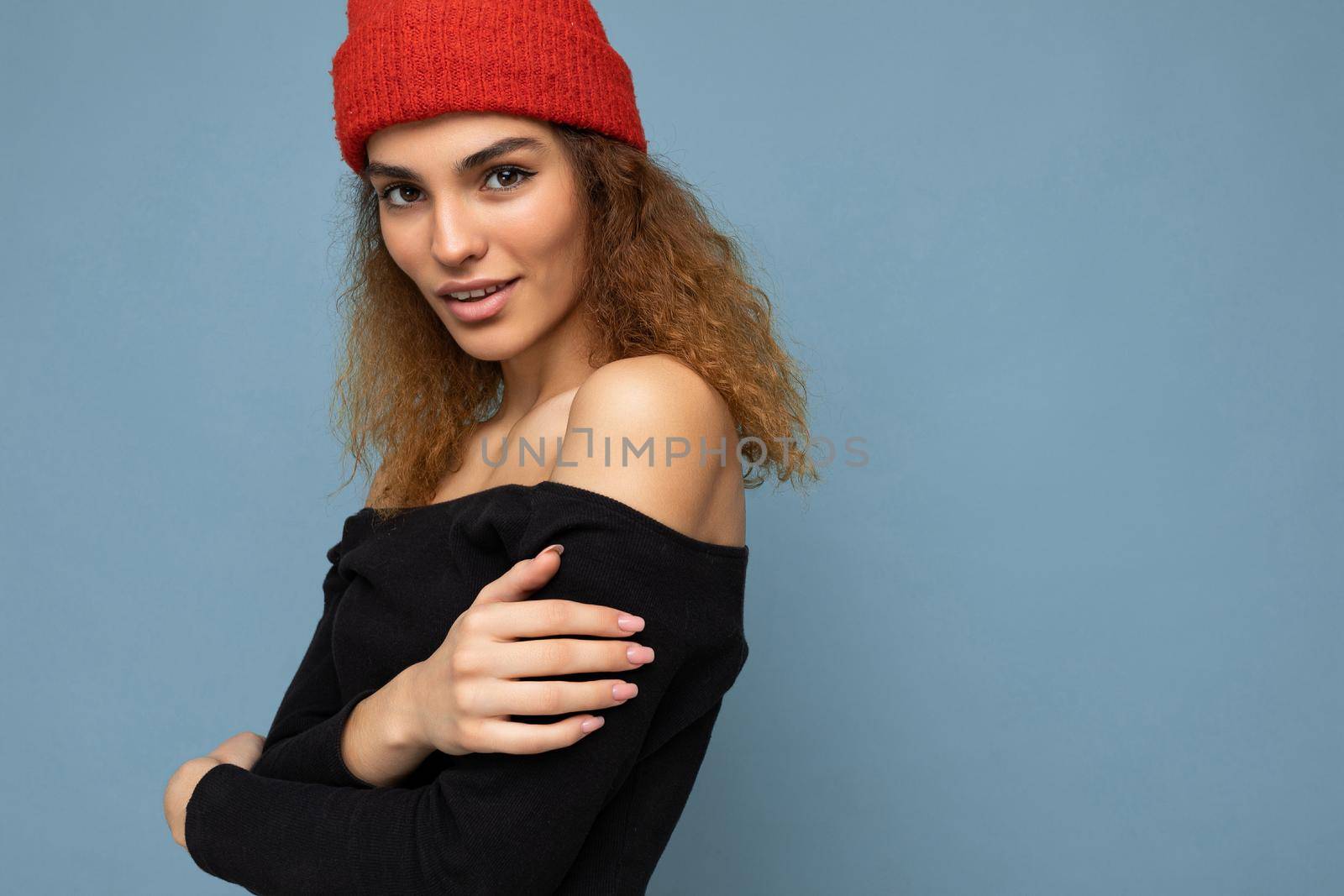 Close-up portrait photo of pretty beautiful attractive cool young curly dark blonde woman standing isolated over blue background wall with copy space for text wearing everyday trendy and stylish black top and red hat.