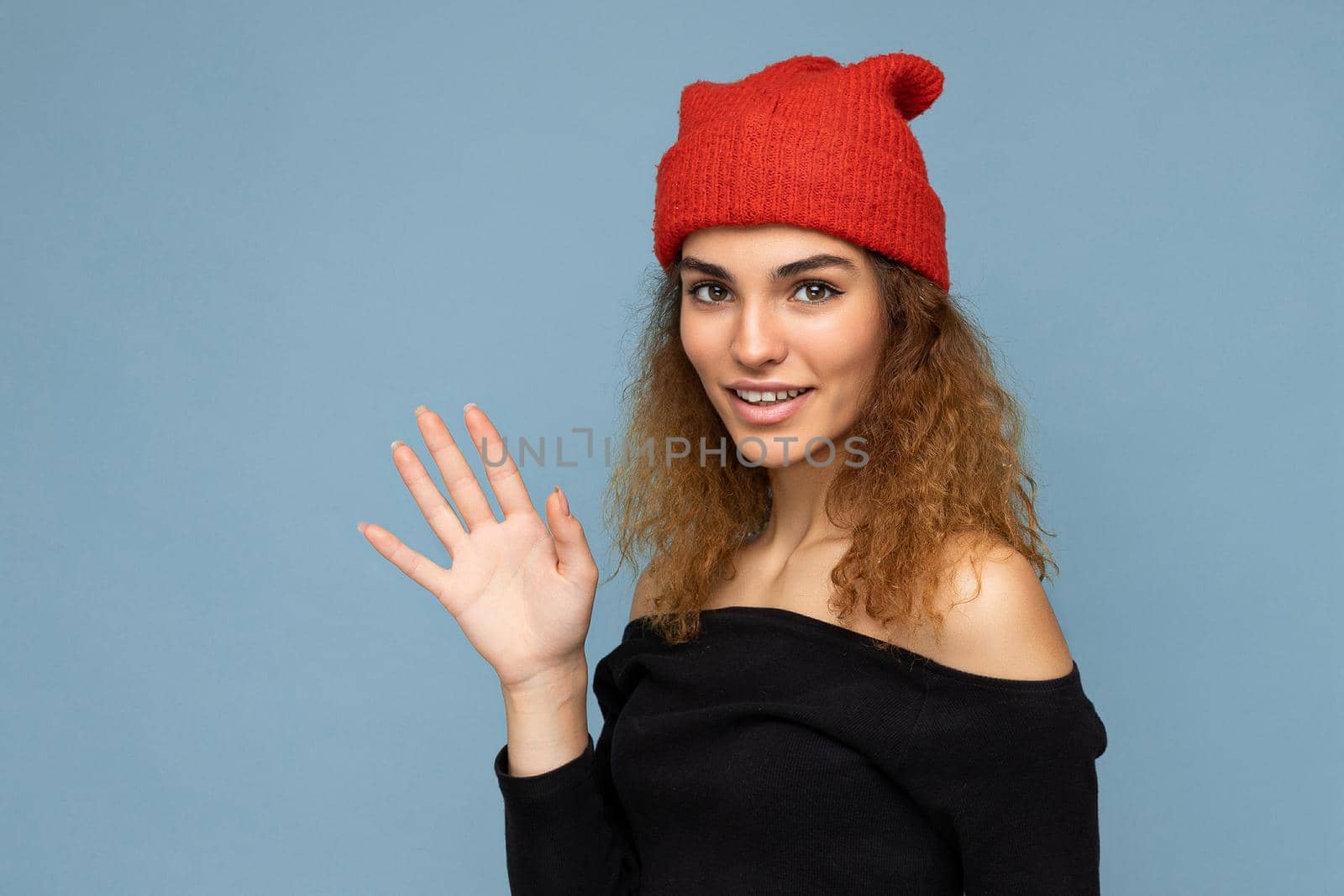 Photo of young positive cute nice brunette woman curly with sincere emotions wearing stylish black crop top and red hat isolated on blue background with copy space and showing palm. Hello concept by TRMK