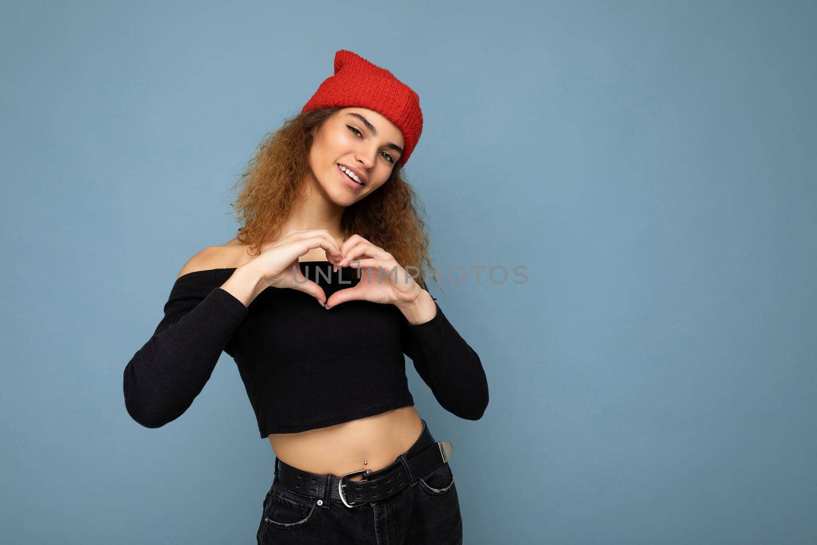 Photo of pretty beautiful attractive chatming smiling happy cool young curly dark blonde woman standing isolated over blue background wall with copy space for text wearing everyday trendy and stylish black top and red hat and showing heart and love gesture with hands.
