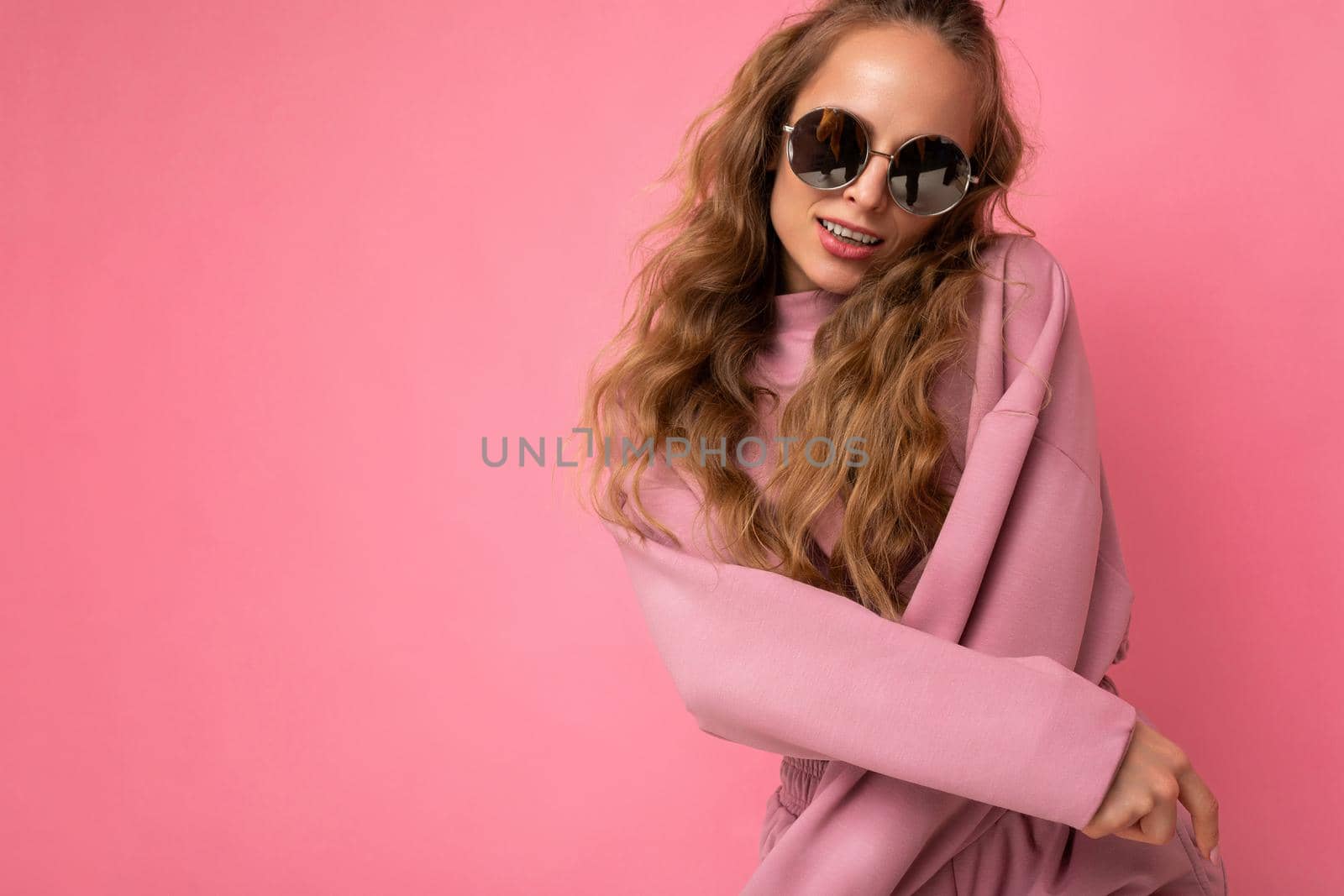 Photo shot of young beautiful attractive cute cool positive curly blonde woman wearing trendy and stylish pink sport suit and sunglasses isolated over pink background with copy space for text.