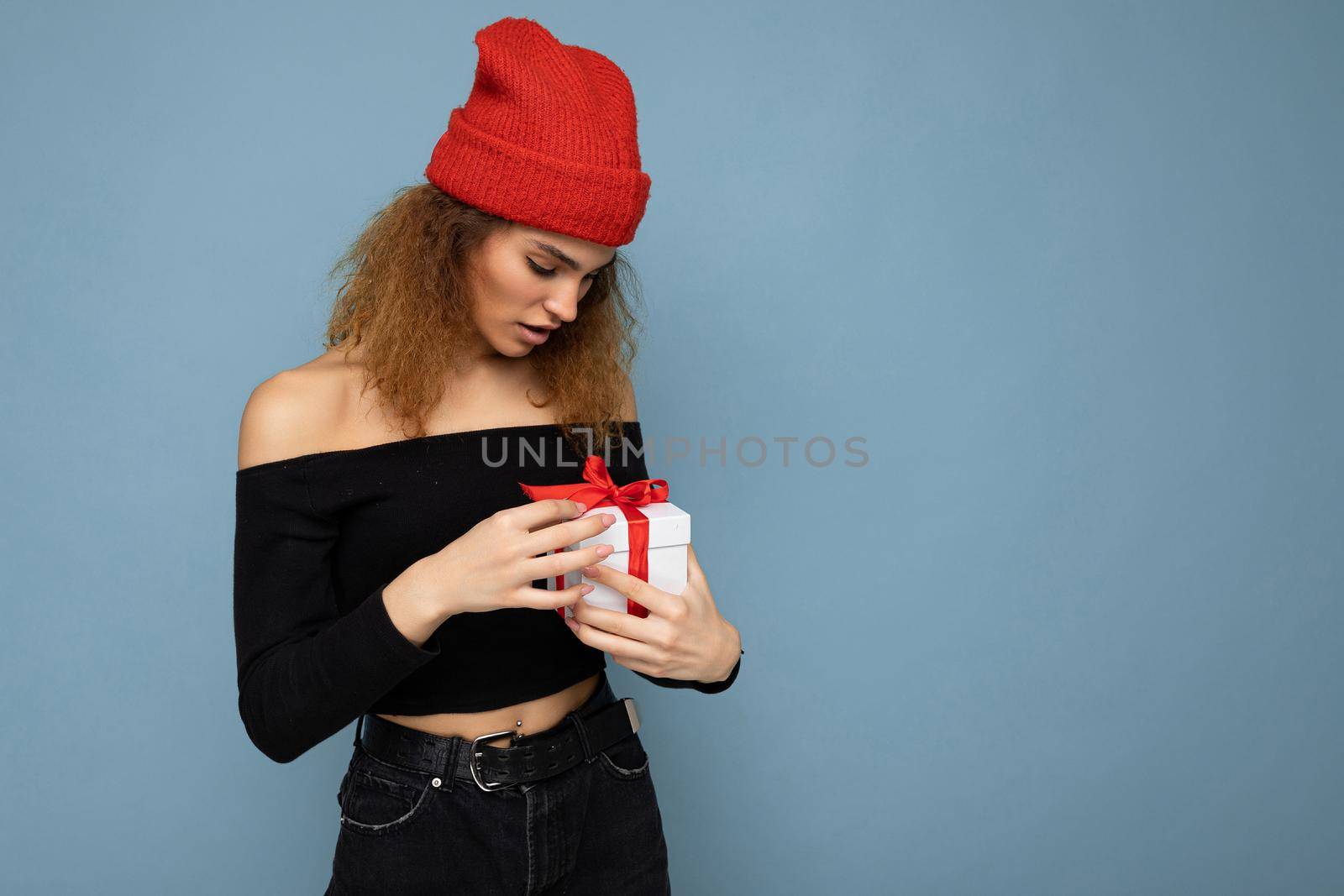 Shot of pretty positive surprised young dark blonde curly woman isolated over blue background wall wearing black crop top and red hat holding gift box looking at surprise box. Copy space, mockup