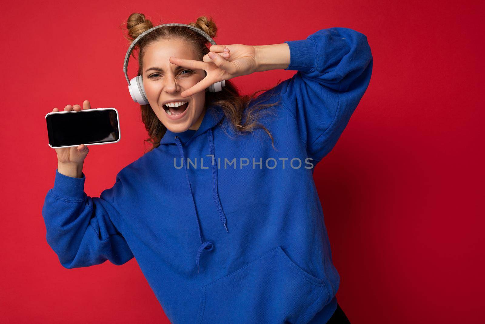 Photo of beautiful positive happy funny young woman wearing stylish blue hoodie isolated over red background with free space for text wearing white bluetooth wireless headphones and listening to music and showing mobile phone with empty screen display for mockup. Peace gesture with two fingers.