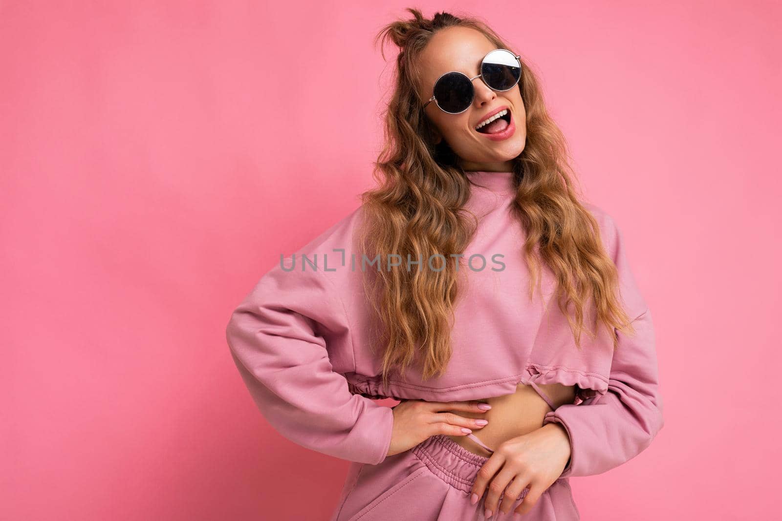 Photo shot of young beautiful attractive cute joyful happy positive laughing curly blonde woman wearing trendy and stylish pink sport suit and sunglasses isolated over pink background with copy space for text.