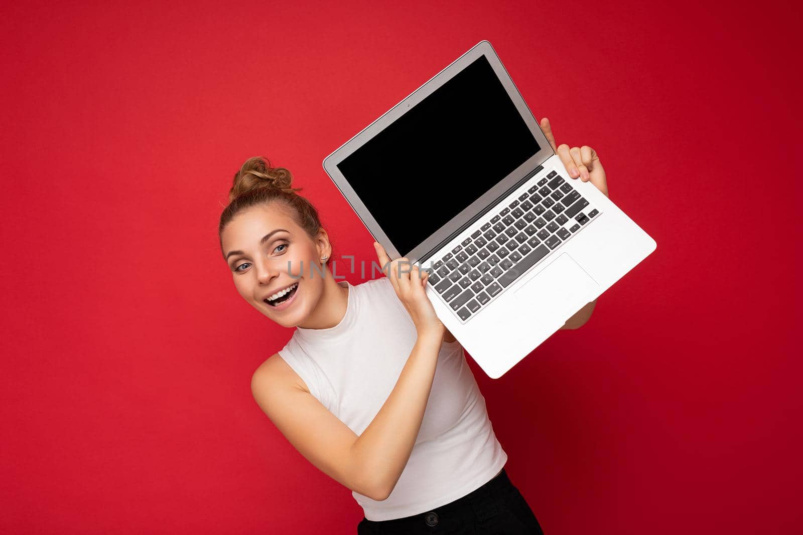 Photo shot of beautiful attractive positive happy smiling young blonde woman isolated over red background wall with empty space for text wearing casual white top and holding laptop computer with black blank display screen for mockup.