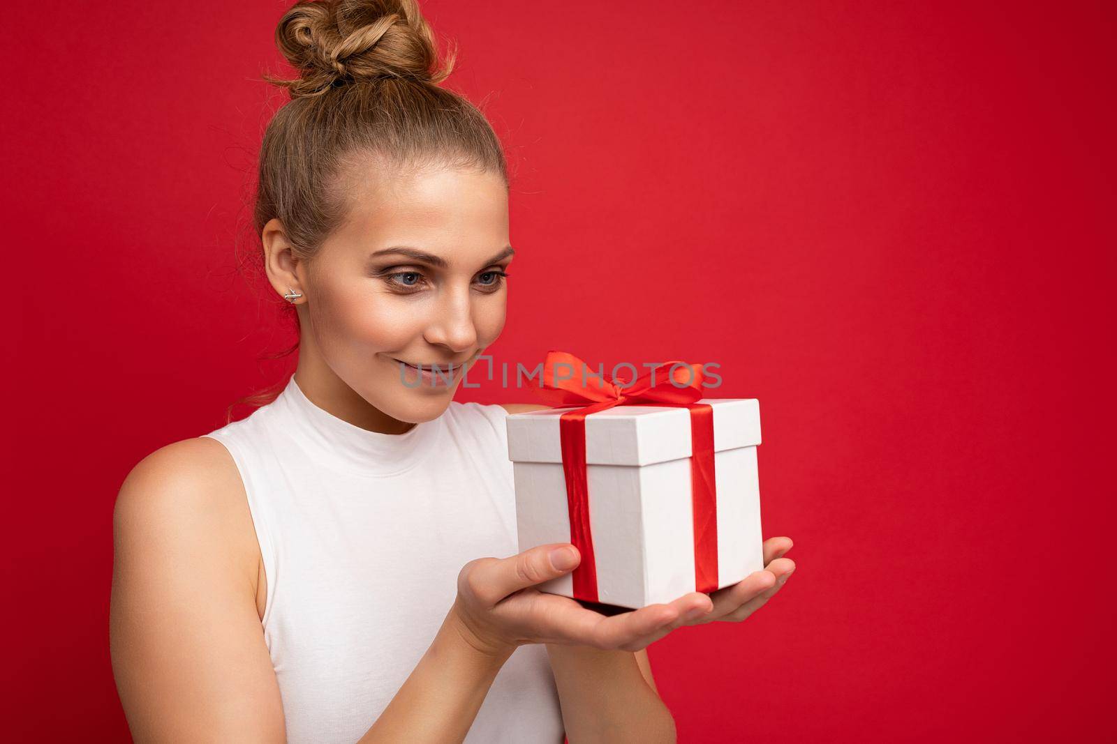 Photo shot of beautiful attractive cute positive smiling thinking young blonde woman isolated over red background wall with copy space for text wearing casual white top holding gift box with red ribbon. New year concept.